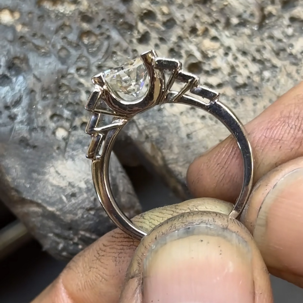 Bague de fiançailles en diamants blancs vue de profil tenue par un artisan joaillier à l'établi